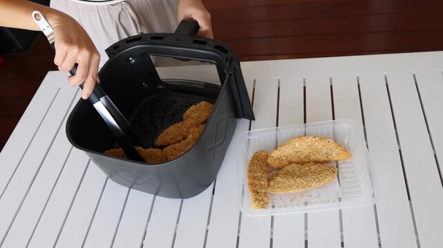 Woman's hands using tongs to place breaded chicken strips into an air fryer basket for cooking.