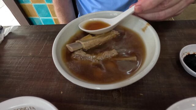 Person eating traditional herbal pork rib soup with spoon and fingers in local restaurant.