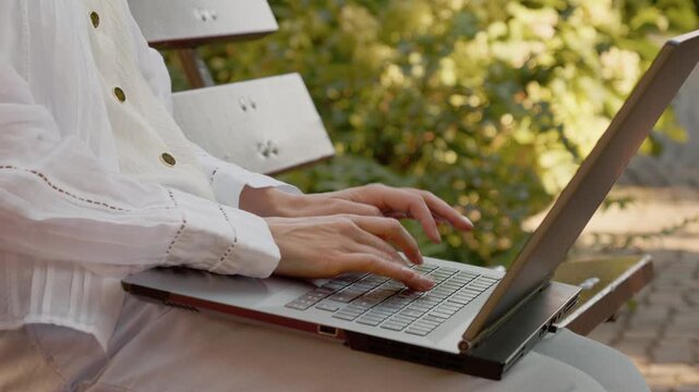 A woman sits on a bench in an outdoor area, typing on a laptop. The setting has green plants around, creating a natural backdrop for her work.