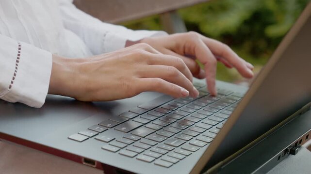 A woman is typing on a laptop while sitting outside. The setting is bright and showcases her hands moving on the keyboard with focus on her task.