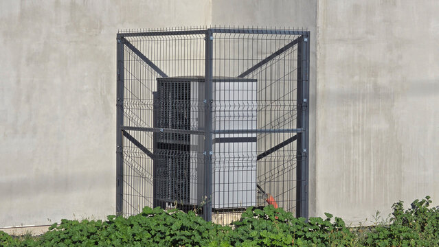 Secure industrial air conditioning unit with protective metal cage fencing and concrete wall backdrop