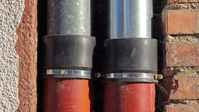 Two large vertical pipes connected by black rubber couplings and hose clamps within a wall recess featuring brick and stucco textures
