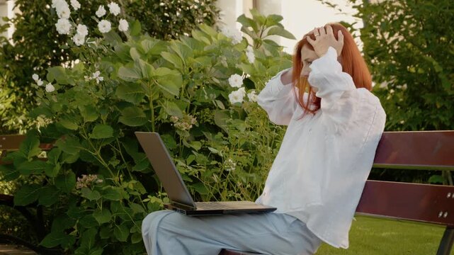 A woman sits on a bench outside, looking at her laptop. She appears sad, clutches her head, and shows clear signs of distress while surrounded by greenery.