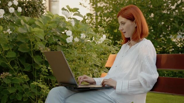 A woman sits on a bench outside, focused on her laptop. She suddenly smiles and raises her hands in joy, celebrating her success.