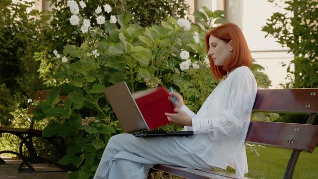 A woman sits on a bench in a garden, using her laptop. She then takes her notebook from the bench and writes some notes before returning to her work.
