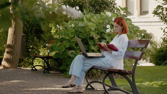 A woman is on an outdoor bench in a garden. She is using a laptop and writing notes in her notebook while looking at the screen.