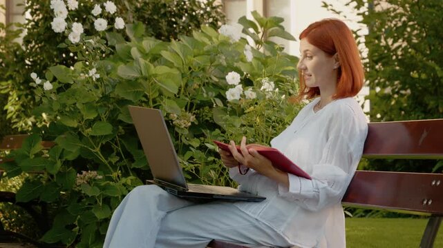 A woman sits on an outdoor bench with her laptop open. She talks on a video call and looks at a notebook she picks up from the bench.