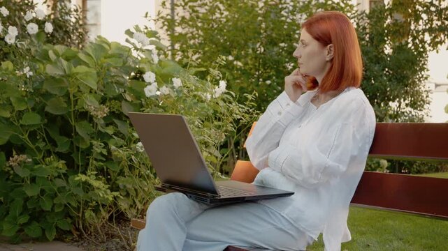 A woman sits on a bench in a garden. She works on her laptop and appears thoughtful. The setting has green plants and flowers, showing a nice outdoor space.