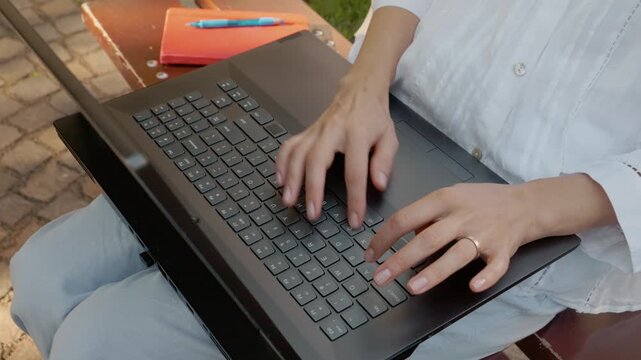 A woman is sitting on an outdoor bench typing on her laptop. She is focusing on her work as she sits comfortably in a natural setting.