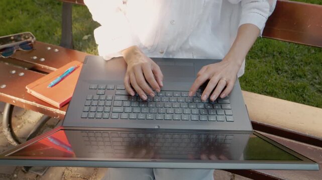 A woman sits on an outdoor bench typing on a laptop. Her hands move quickly over the keyboard as she engages in her work in a relaxed environment.