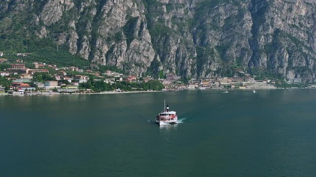 Cinematic rotational 4k zoom drone shot of a large classic motor vessel underway on Lake Garda with dramatic mountains and an Italian village in the background