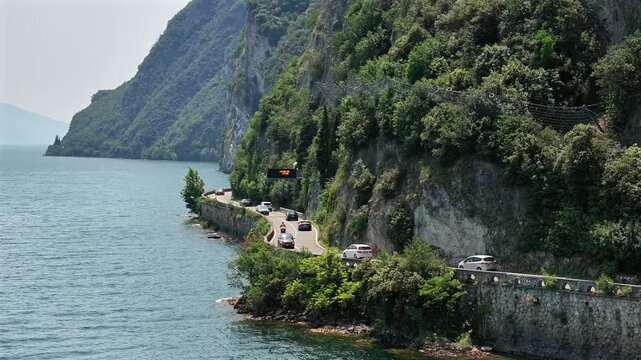 Cinematic drone scene of a road next to Lake Garda in Italy with dramatic, steep cliffs and rockfall netting protecting cars and drivers as they commute along the winding road into a tunnel