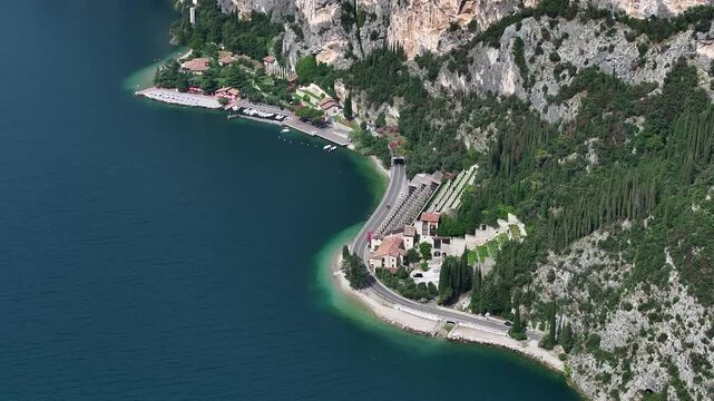 High altitude, cinematic 4k drone shot of dramatic cliffs at the edge of Lake Garda with winding road following the coastline and beautiful sandy beaches near the quaint village of Tignale