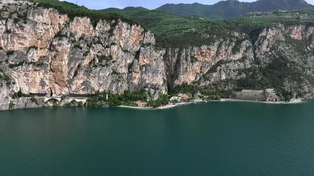 Wide 4k cinematic drone shot of massive, sheer cliffs and mountains next to Lake Garda with Tignale beach and quaint Italian villages in the European summer