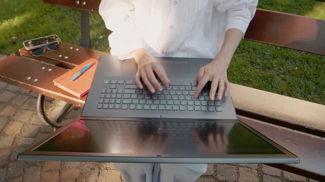 A female student types on a laptop while sitting on a bench outside. She has a notebook and sunglasses next to her. It is a sunny day and the grass is green.