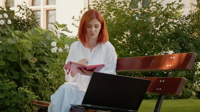 A female student sits on a bench with a small notebook while she chats on a video call using her laptop. She seems happy and engaged during her conversation.