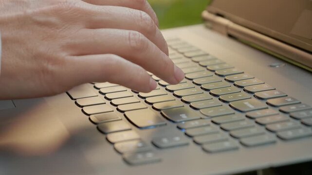 A close-up shot captures a woman's fingers moving across a laptop keyboard. The fingers are typing in the bright outdoor light.
