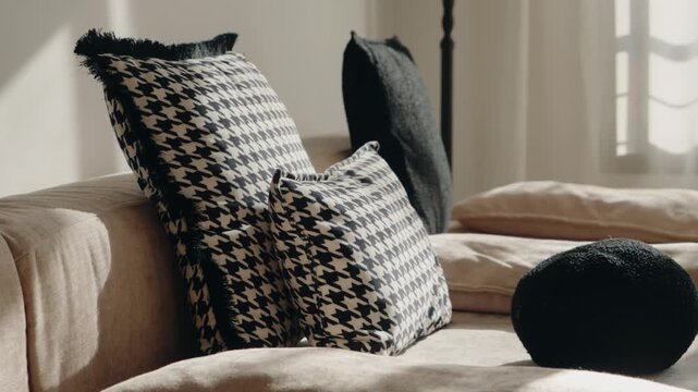Sunlight streams into a minimally decorated room, illuminating throw pillows arranged on a couch. The focus is on the textures and patterns, including houndstooth and a fuzzy black pillow.