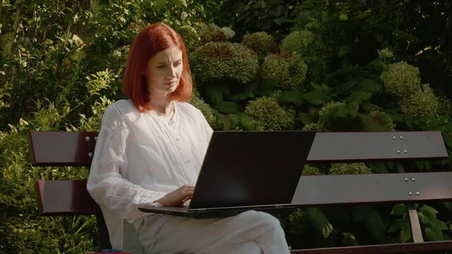 A female student sits on a bench outside in a garden. She is focused on her laptop while surrounded by plants and flowers during the day.