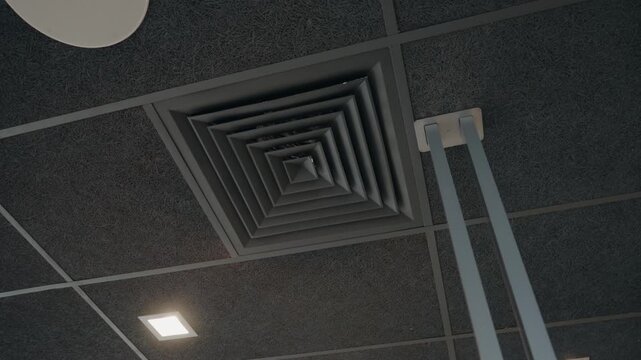 Looking up at a modern office ceiling with black acoustic tiles, square LED lights, a circular speaker, and a square HVAC vent. Hanging lights descend from thin gray poles,