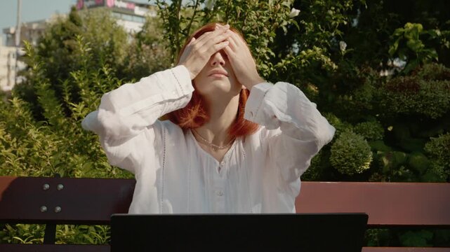 A female student sits on a bench outside in a garden. She looks at her laptop and shows sadness. She raises her hands and places them on her head.