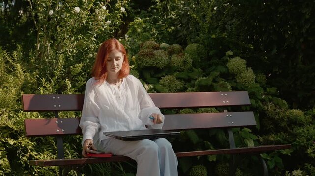 A female student sits on a bench in a garden working on her laptop. After some time, she closes it and walks away from the bench.