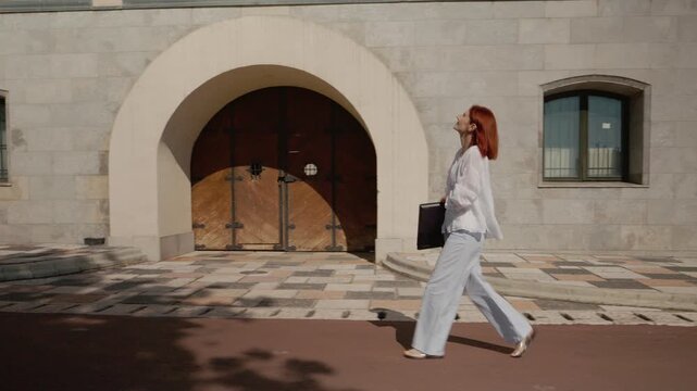 A female student walks along a path next to a university building while carrying a laptop in her hand. She moves steadily on a clear day.
