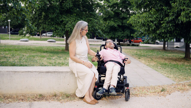 Caregiver sitting beside person with spina bifida in motorized wheelchair, providing support and companionship in an urban park