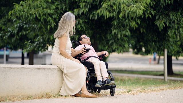 Adult woman supporting a man with spina bifida in a powered wheelchair, enjoying time together in a park