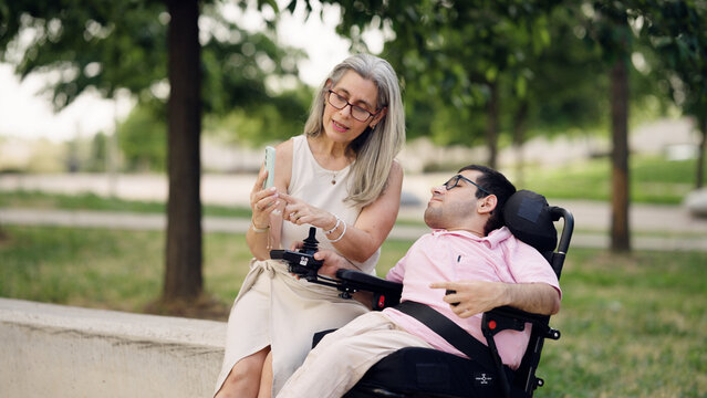 Caregiver assisting a man with spina bifida in a wheelchair outdoors, sharing content on a smartphone in a park