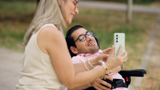 Caregiver assisting a man with spina bifida in a wheelchair, both smiling while looking at a smartphone outdoors