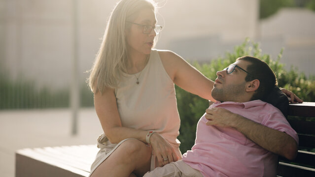 Caregiver sitting with a man with physical disability on a bench outdoors, offering support and companionship