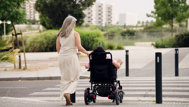 Caregiver pushing an adult with spina bifida in an electric wheelchair, navigating an accessible urban park pathway
