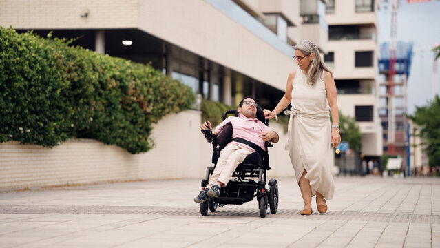 Man with physical disability using electric wheelchair on city street, assisted by a woman. Depicting inclusion and mobility