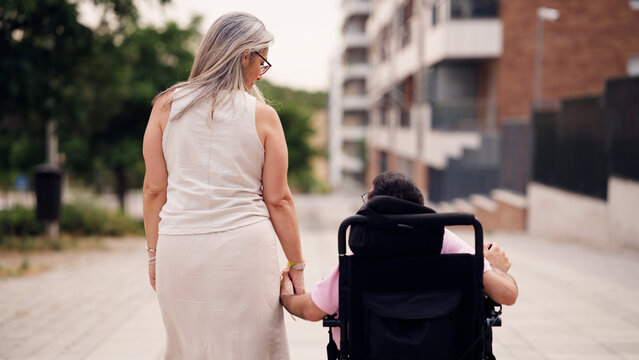 Caregiver holding hand of person with physical disability in wheelchair, navigating urban outdoor environment together
