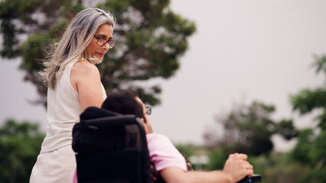 Woman assisting an adult male with spina bifida in an electric wheelchair outdoors, providing support and companionship