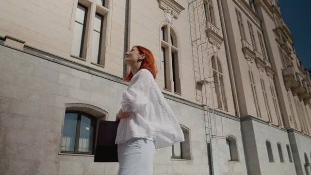 A female student walks along a path next to a university building holding a laptop. She turns around and looks at her surroundings during the day.