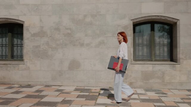 A female student walks along a path next to a university building. She carries a heavy laptop while moving toward her destination.