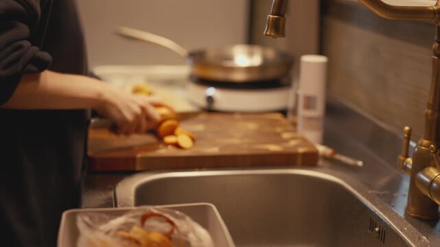 Selective focus close-up on brass faucet and kitchen sink with woman slicing peeled sweet potato with knife on wooden board in blurred background. Food prep process in cozy warm home kitchen.