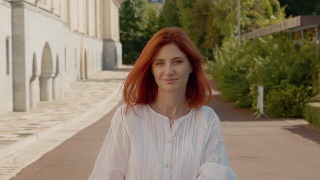 A red-haired woman follows the camera with her gaze while walking in a park on a sunny day. There are trees and people in the background enjoying their time outside.