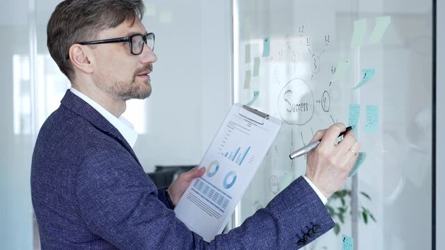 Business man evaluating success strategy roadmap on a glass wall, analyzing project development and financial data charts on a clipboard in a modern office
