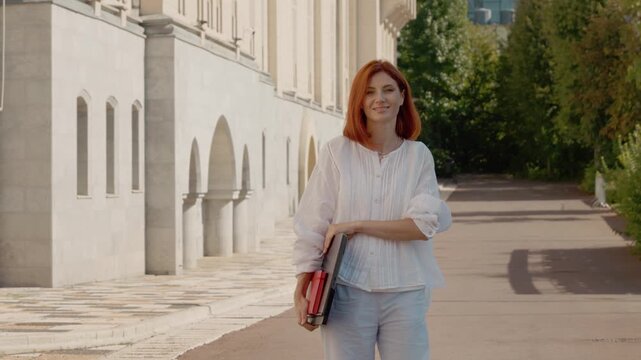 A red-haired woman turns and looks at the camera while walking through a park. She holds a laptop in her hands as she walks along the path.