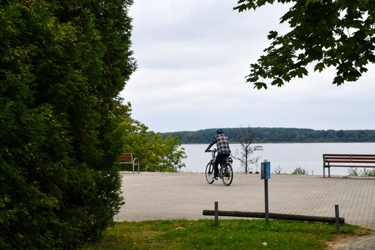 Radfahrer an einer Uferpromenade mit weitem Blick &uuml;ber einen See. Fahrradtour, Freizeit, Natur, Erholung, Radweg, Bewegung, Aktivurlaub und sommerliche Landschaft am Wasser.