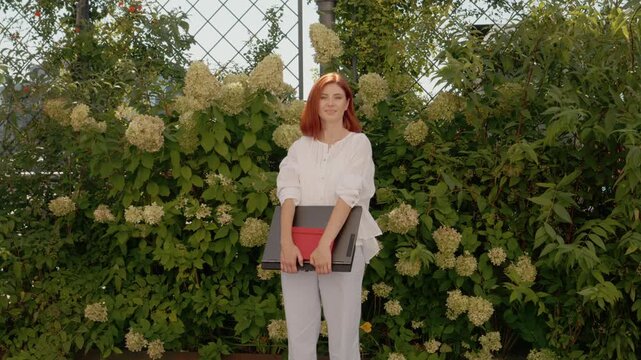 A redhead woman looks at the camera while standing outside, holding a laptop. She is surrounded by green plants and white flowers on a sunny day.