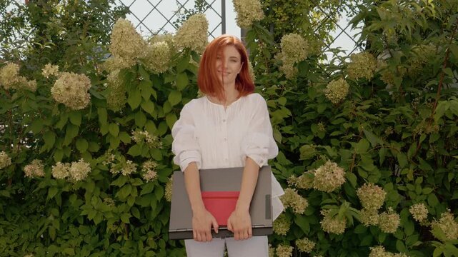 A redhead woman stands outdoors holding a laptop. She looks directly at the camera while surrounded by green plants and flowers on a sunny day.