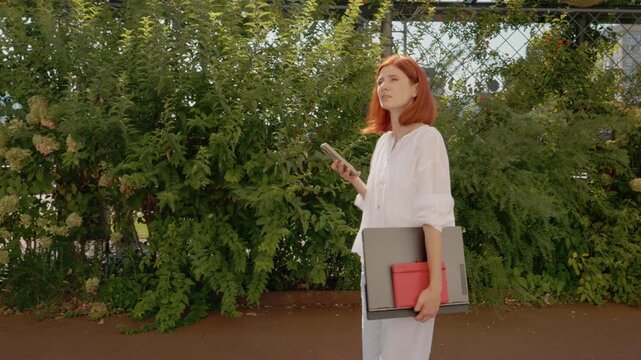 A female student is walking in a garden area, holding a laptop in one hand and a phone in the other, looking unsure of where to go next.