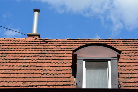 A clay tile roof of a residential house featuring a modern metal chimney liner and an attic dormer window under a blue sky.