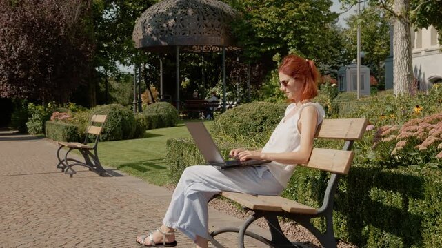 A student sits on a bench in a park typing on a laptop. The area features bushes and flowers. It appears to be a sunny day.