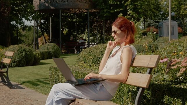 A student sits on a park bench in a garden, typing on a laptop. The sun shines and the area is green with plants and flowers around.