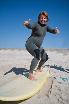 Happy plus size woman in wetsuit balancing on surfboard at beach showing thumbs up